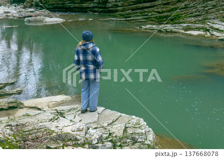 young woman in a plaid jacket is standing on a rock by a river. 137668078