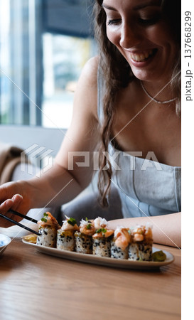 A beautiful girl eats Japanese sushi rolls with chopsticks, smiling with pleasure A beautiful girl eats Japanese sushi rolls with chopsticks, smiling with pleasure 137668299