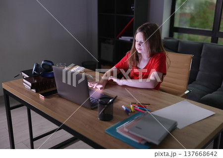 Young girl immersed in learning, typing on laptop at home desk in evening light with vibrant creativity around her 137668642