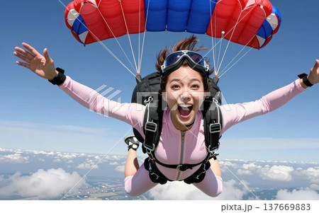 Thrilling skydiving portrait of a joyful young Asian woman in jumpsuit and goggles, soaring with arms outstretched under a colorful parachute amid blue skies and clouds. 137669883