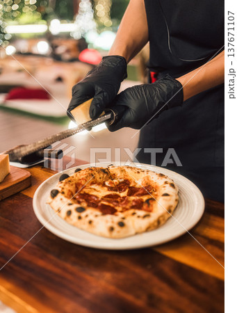Hand chef grating parmesan cheese over atrisan pepperoni pizza on wooden table Hand chef grating parmesan cheese over atrisan pepperoni pizza on wooden table 137671107