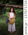 Little girl with long curly hair holding a woven straw bag with purple flowers in a forest path 137671726