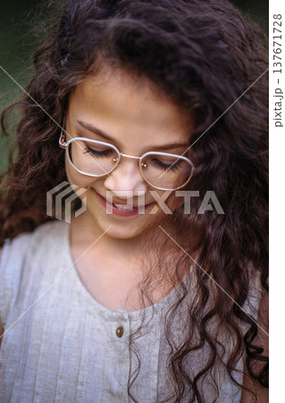 Close up portrait of a smiling young girl with long curly hair and glasses looking down 137671728