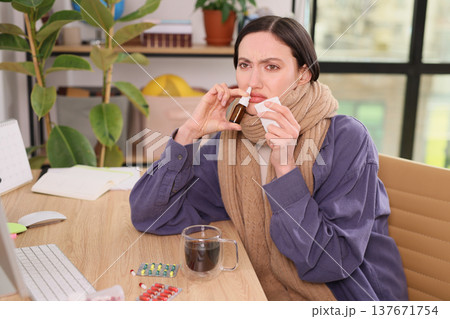 Sick woman sits at desk taking medicine and sipping coffee while wrapped in a cozy scarf during a cold morning at home 137671754