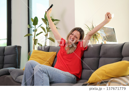 Joyful moment captured as a woman celebrates success on a cozy sofa in a sunny living room filled with plants and warmth 137671779