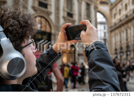 Milan, italy, april 1, 2024. Young man wearing headphones and glasses taking vertical smartphone video or picture of classical architecture inside famous milan gallery Milan, italy, april 1, 2024. Young man wearing headphones and glasses taking vertical smartphone video or picture of classical architecture inside famous milan gallery 137672331