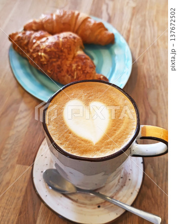 Cup of latte with heart-shaped foam art served with fresh croissant on a rustic wooden table. 137672502