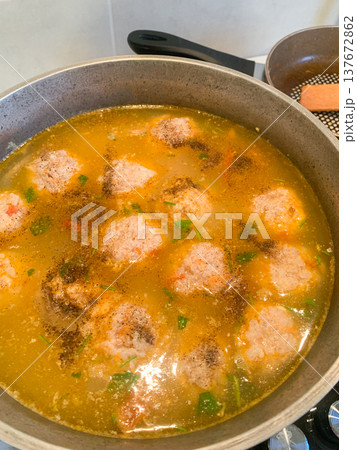 Meatballs are cooking in a pot of broth on the stove. The broth includes spices and vegetables. This shows meal preparation in a home kitchen 137672862