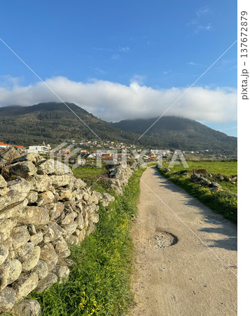 Oia, Galicia, Spain - March 27, 2025: Camino de Santiago rural path leading toward village houses, green hills with forested mountains. Camino de Santiago route landscape, rural village scenery Oia, Galicia, Spain - March 27, 2025: Camino de Santiago rural path leading toward village houses, green hills with forested mountains. Camino de Santiago route landscape, rural village scenery 137672879