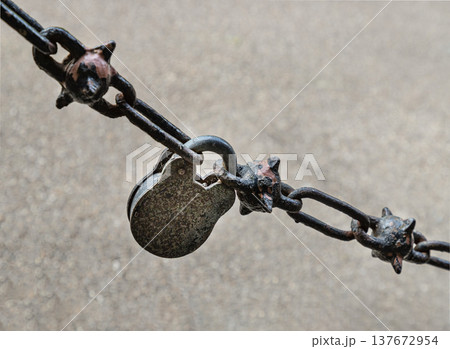 A weathered, vintage-style padlock serves as the central link in a heavy spiked chain. This Gothic-inspired hardware, featuring ornate four-pointed studs on every other link A weathered, vintage-style padlock serves as the central link in a heavy spiked chain. This Gothic-inspired hardware, featuring ornate four-pointed studs on every other link 137672954