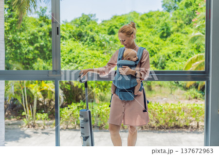 Woman vacuuming a room while carrying her baby in an ergonomic carrier. Modern motherhood, multitasking parent, home chores with infant, balance of childcare and housework concept. 137672963