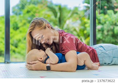 Mother and baby lying on a soft play mat at home, playing, talking and sharing a joyful moment together. Authentic family lifestyle, early childhood development and emotional bonding. Parenting and Mother and baby lying on a soft play mat at home, playing, talking and sharing a joyful moment together. Authentic family lifestyle, early childhood development and emotional bonding. Parenting and 137673622