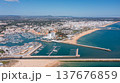 People visit Vilamoura marina in Portugal's Algarve region, enjoying boats and beach while the city skyline stands tall in the distance under clear blue sky Aerial 137676859