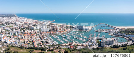 People enjoy tourism in Vilamoura as they visit the marina, observe boats, and take in views of the surrounding coastal landscape and city buildings Aerial panorama People enjoy tourism in Vilamoura as they visit the marina, observe boats, and take in views of the surrounding coastal landscape and city buildings Aerial panorama 137676861
