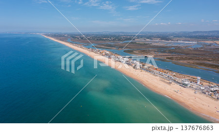 View of Ria Formosa and Praia de Faro taken from a drone. The coastline features sand and water, with nearby buildings and greenery visible 137676863