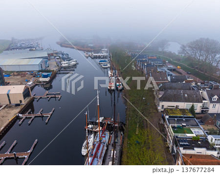 Aerial view of Muiden harbor in the fog, featuring traditional Dutch sailboats, a marina, and the silhouette of Muiderslot castle in the misty background. 137677284