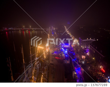 Aerial night view of the Kampen Christmas market along the IJssel river. The scene features historic sailing ships with festive lights, a crowded quay, and the illuminated city bridge in the mist. 137677289