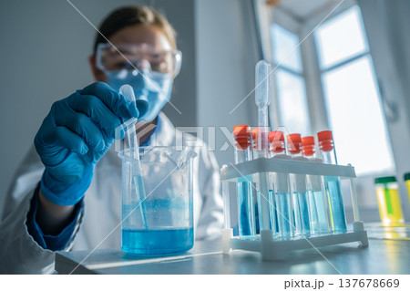 A researcher wearing gloves and safety gear uses a pipette to transfer blue solution into a beaker in a modern lab. Sunlight streams through the window, illuminating scientific equipment and glassware 137678669