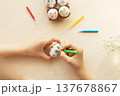 Children's hands use green marker to color simple flower designs on white eggs on table, with other eggs in tray and markers nearby, Easter, handmade 137678867