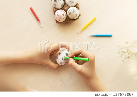 Children's hands use green marker to color simple flower designs on white eggs on table, with other eggs in tray and markers nearby, Easter, handmade 137678867
