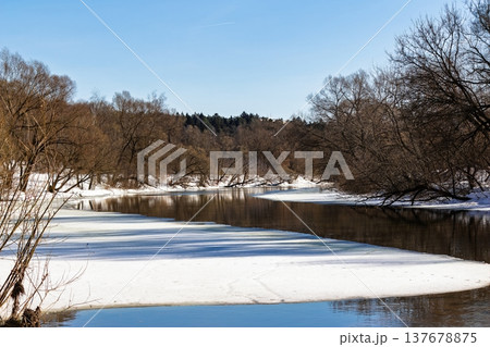 Melting river ice with open dark water reflecting bare branches under a bright blue spring sky 137678875