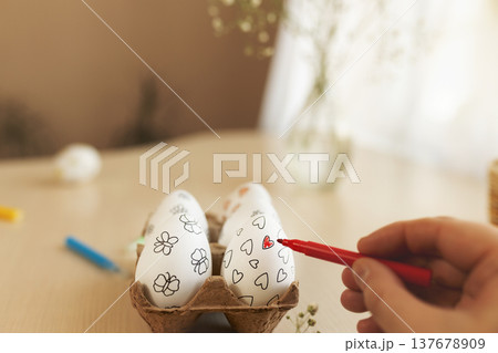 Close-up of person decorating Easter eggs with red marker. White eggs with hand-drawn hearts and flowers placed in an egg carton on table. Creative Easter preparation, DIY holiday decoration 137678909