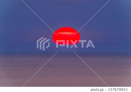 水平線に浮かぶ巨大な赤いだるま夕日と静かな海の幻想的な風景 137678931