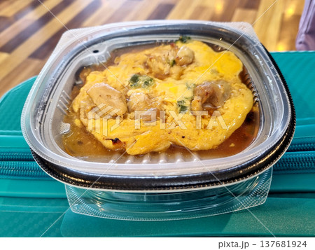 Japanese Oyakodon take-out meal with chicken and egg in a clear plastic container. Authentic bento box dish with savory sauce and green onions, resting on a travel bag in a modern public setting. 137681924