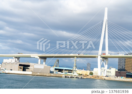 Large white cable-stayed bridge spanning over a harbor with industrial port buildings and cranes in the background. Modern infrastructure and maritime transport scene under a cloudy sky. Large white cable-stayed bridge spanning over a harbor with industrial port buildings and cranes in the background. Modern infrastructure and maritime transport scene under a cloudy sky. 137681936