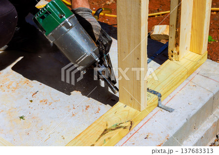 Construction worker uses nail gun to fasten wooden frame on building site Construction worker uses nail gun to fasten wooden frame on building site 137683313