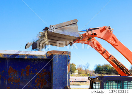 Construction vehicle unloads material into blue dumpster under clear sky at worksite Construction vehicle unloads material into blue dumpster under clear sky at worksite 137683332