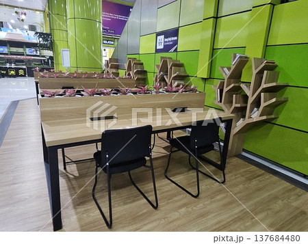 A modern co-working space at a station featuring wooden desks with black chairs, unique tree-shaped bookshelves, and small plants set against a vibrant green wall with integrated power outlets. A modern co-working space at a station featuring wooden desks with black chairs, unique tree-shaped bookshelves, and small plants set against a vibrant green wall with integrated power outlets. 137684480
