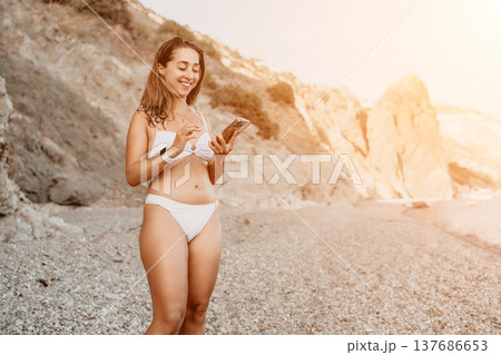 Woman, beach, smartphone. Happy young woman smiling, using phone on a sunny pebble beach with cliffs. 137686653