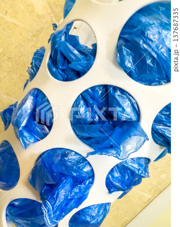 White plastic bag dispenser filled with blue plastic bags, mounted on a tiled wall in a kitchen or utility area 137687335