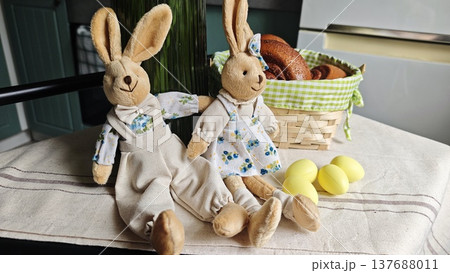 Plush bunny dolls dressed in floral outfits sit beside a wicker basket filled with baked goods and yellow Easter eggs on a table with a linen cloth Plush bunny dolls dressed in floral outfits sit beside a wicker basket filled with baked goods and yellow Easter eggs on a table with a linen cloth 137688011