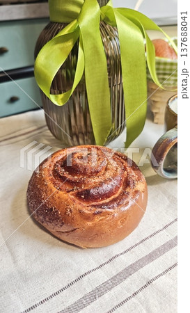 Freshly baked cinnamon roll placed on a linen tablecloth beside a glass jar tied with a green ribbon and a ceramic mug in a cozy kitchen setting Freshly baked cinnamon roll placed on a linen tablecloth beside a glass jar tied with a green ribbon and a ceramic mug in a cozy kitchen setting 137688041