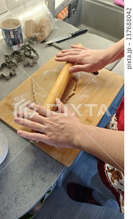 Hands of a person rolling out dough with a wooden rolling pin on a wooden cutting board in a kitchen, cookie cutters and utensils visible in the background 137688042