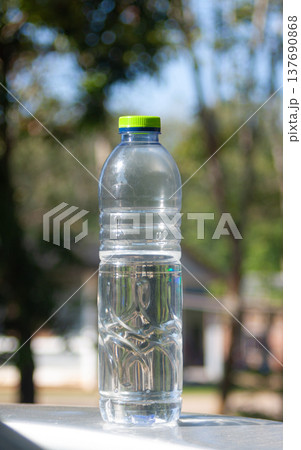 A cold plastic water bottle with condensation stands outdoors, catching sunlight against green blur. 137690868