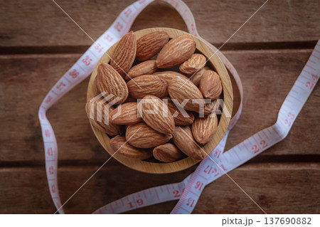 Almonds in a wooden cup on a wood plate with a measuring tape, symbolizing healthy diet control. Almonds in a wooden cup on a wood plate with a measuring tape, symbolizing healthy diet control. 137690882
