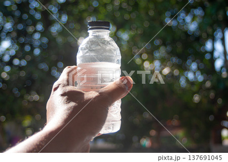 A hand holds a cold plastic water bottle outdoors, glowing in sunlight with soft green bokeh lit sun 137691045