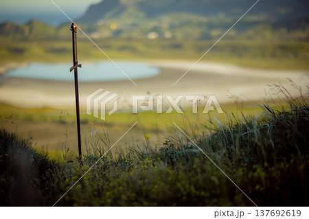 A medieval iron sword is stuck into the ground against a backdrop of mountains and the sea 137692619