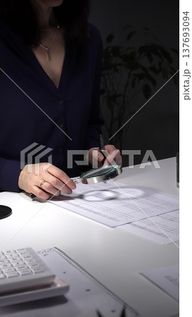 Person checking financial documents and numbers with a magnifying glass, analyzing business data for budgeting and taxes at night. Audit and taxes in business 137693094