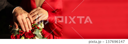 Close-up of a bride and groom's hands with wedding rings on a red rose bouquet. Wedding couple in red dress and dark suit, celebrating marriage. Large red copy space background Close-up of a bride and groom's hands with wedding rings on a red rose bouquet. Wedding couple in red dress and dark suit, celebrating marriage. Large red copy space background 137696147