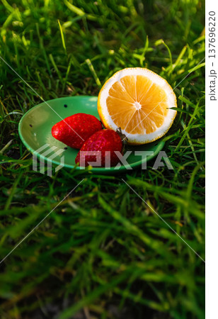 Defocus close-up saucer with lemon and two strawberry standing on deep green grass. Blurred nature background. Bright summer fruits outside. Sunshine. Vitamin c. Out of focus 137696220