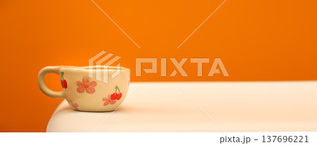 Minimalist still life with a ceramic cup on a white table. Beige handmade mug decorated with pink flowers and cherries against a solid orange background. Cozy morning and copy space concept 137696221