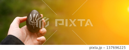 Patriotic Ukrainian Easter concept. A hand holding a dark Easter egg with a hand-drawn white Trident, Tryzub, the national symbol of Ukraine. background with bright sun flare and copy space Patriotic Ukrainian Easter concept. A hand holding a dark Easter egg with a hand-drawn white Trident, Tryzub, the national symbol of Ukraine. background with bright sun flare and copy space 137696225