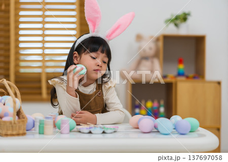 preschool child girl wearing bunny ears holding colorful egg while celebrating on easter day on table at home preschool child girl wearing bunny ears holding colorful egg while celebrating on easter day on table at home 137697058