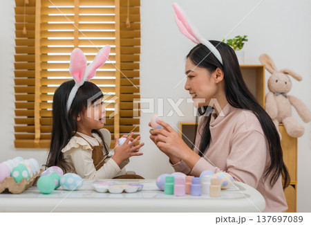 mother and preschool child girl wearing bunny ears holding colorful egg while celebrating together at home on easter day. happy family holiday activity concept mother and preschool child girl wearing bunny ears holding colorful egg while celebrating together at home on easter day. happy family holiday activity concept 137697089