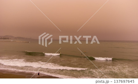 Surreal Beach Scene: Spanish Shoreline under Orange Sky from Moroccan Sandstorm. Captivating moment with waves, surfers, and swimmers enveloped in dramatic natural hues.Spain in sandstorm from Moroco 137697645