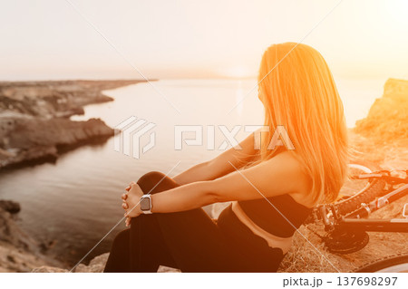 Woman sunset bicycle resting on cliff overlooking ocean after bike ride during golden hour Woman sunset bicycle resting on cliff overlooking ocean after bike ride during golden hour 137698297